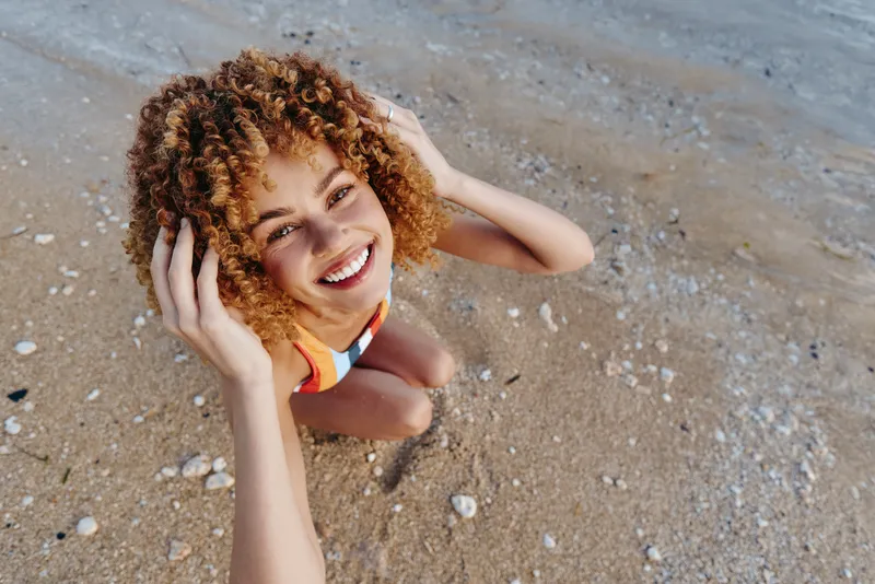 Young woman with curly hair smiles on a sandy beach, wearing a swimsuit, photographed from above as she enjoys a sunny day by the water. Her cheerful expression and relaxed pose convey carefree