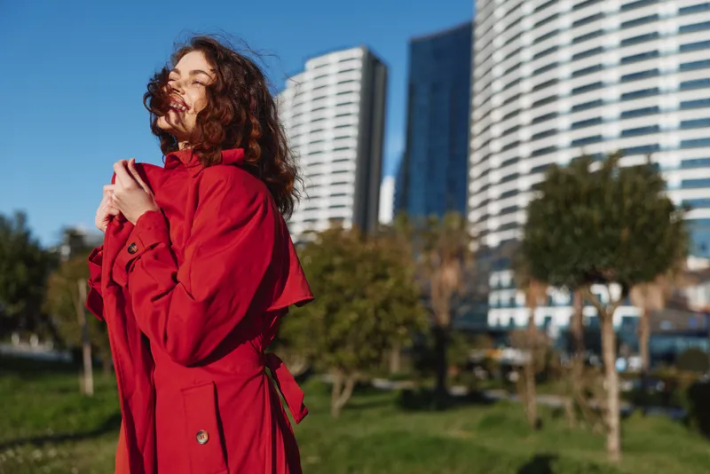 A young woman in a red coat enjoys sunshine outdoors with eyes closed, standing in a city park with modern buildings in the background on a clear day.