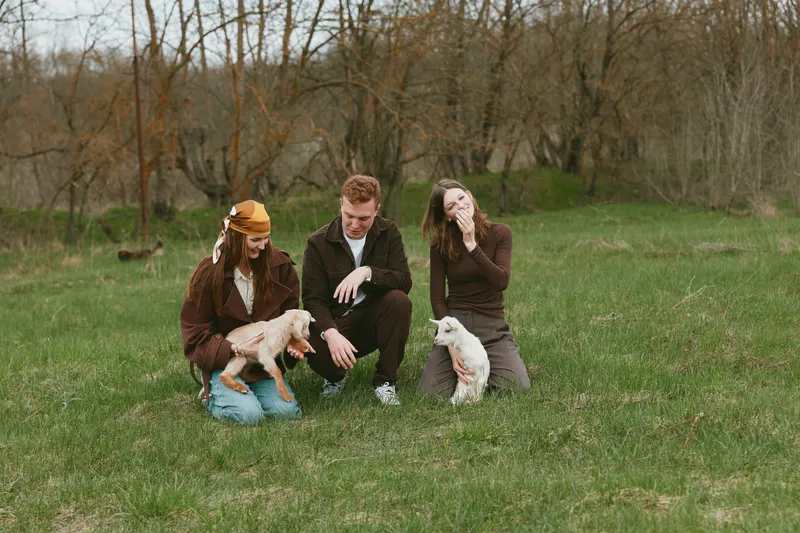 Lifestyle scene with three friends in a grassy park, interacting with a playful goat kid, warm natural light, relaxed smiles, candid moment of companionship and outdoor simple joy