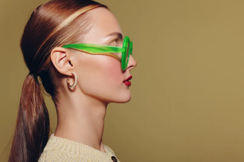 Profile of a young woman with sleek hair tied back wearing green sunglasses and hoop earrings on beige background. Concept of fashion, style, and modern accessories.