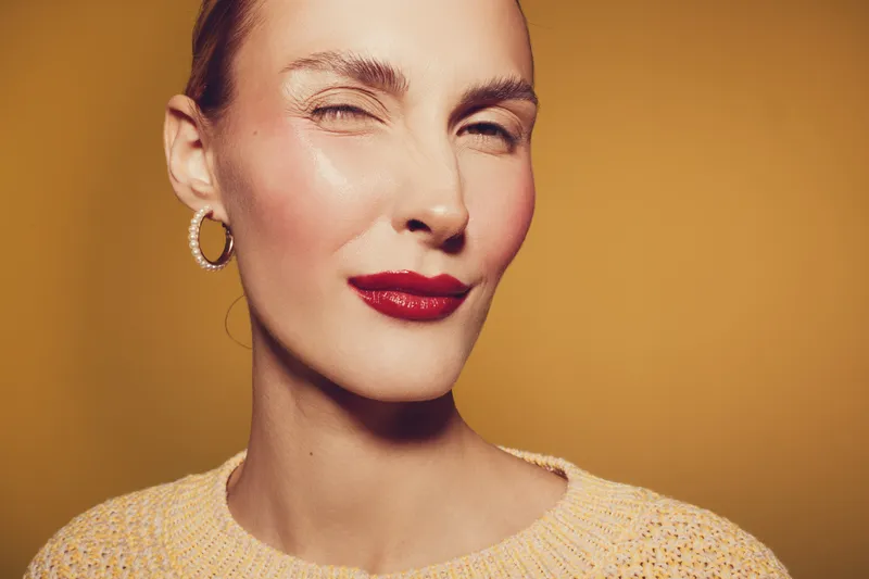 Close-up portrait of confident woman with red lipstick winking, wearing hoop earrings and yellow sweater. Warm studio lighting on orange background creates bold and stylish mood.
