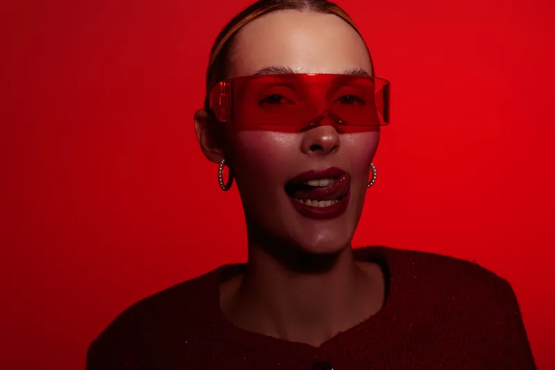 Young woman wearing red sunglasses and dark lipstick poses with tongue out against red background. Studio portrait with dramatic lighting and bold fashion style concept.