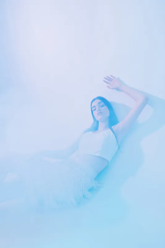 Young woman in white fashion outfit lies on floor with eyes closed. Soft blue lighting creates dreamy beauty concept for fashion and wellness.