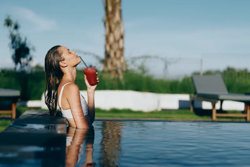 Young woman relaxes in a pool, sipping a red drink from a glass, enjoying a sunny day by the water, casual and carefree in bright light.