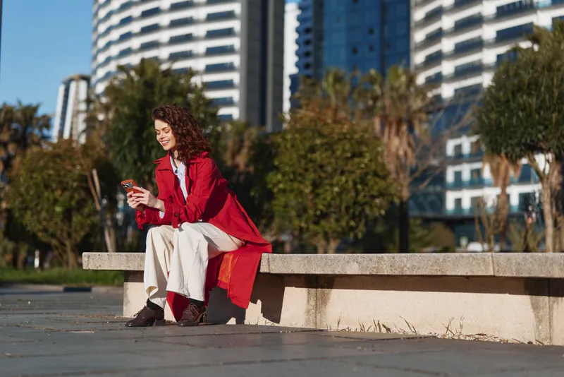 Young woman with curly hair wearing a red coat sits on a concrete bench in an urban park while using her smartphone and smiling happily.