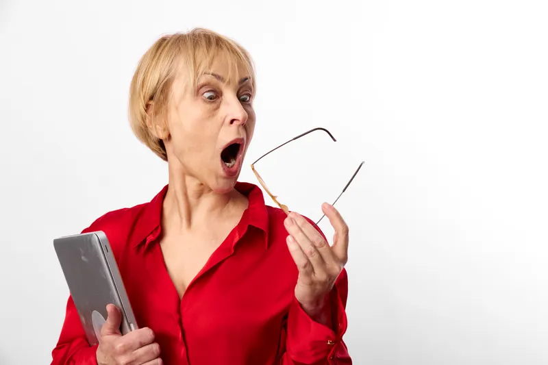 Elderly woman shows surprise indoors holding a smartphone and glasses, red shirt, shocked face, white background, hands raised, modern communication moment