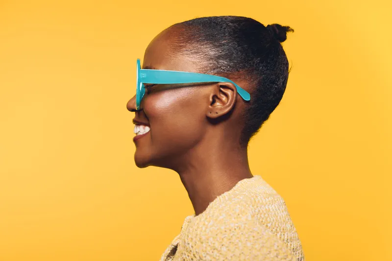 Profile of a smiling young African woman with dark skin wearing blue sunglasses and a beige sweater on yellow background. Concept of fashion, lifestyle, and summer accessories.