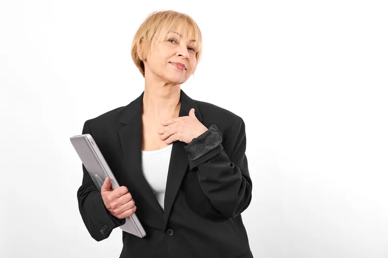 Woman with tablet in business portrait, smiling senior woman in black blazer, white background studio lighting, hand on chest, calm confident mood, technology enabled career