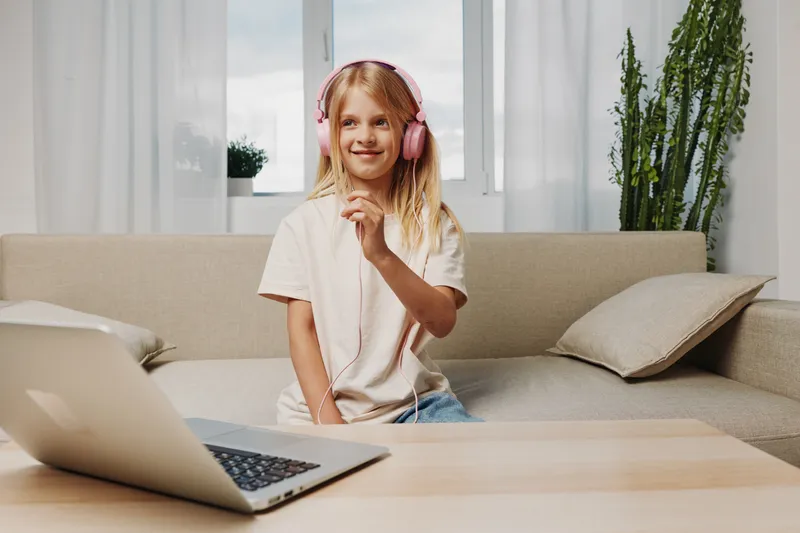Smiling blonde girl with pink headphones listens to music near laptop in cozy living room with modern sofa and houseplants.