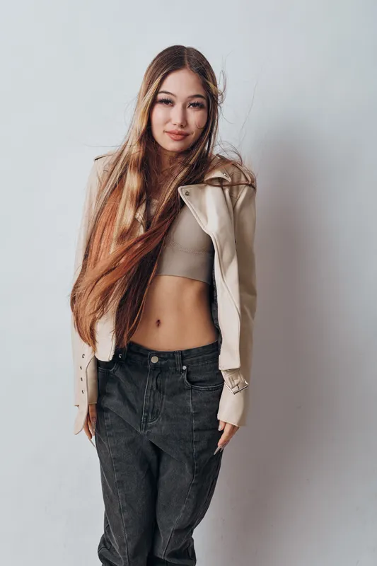 Young woman with long hair wearing casual outfit including crop top and jacket posing against light background. Fashion model with natural look and confident expression indoors.