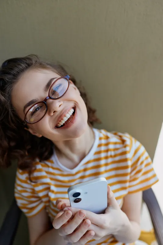 A cheerful woman in a striped shirt with glasses sits indoors, smiling at a smartphone. A candid moment of home life, tech use, and personal connection.