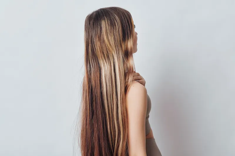 Woman with long straight hair and light blonde highlights in studio. Back view of female model with smooth skin against neutral background for hairstyle and fashion.