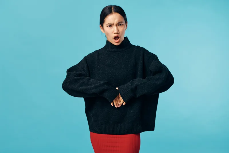 Woman in black sweater and red skirt showing disgust or frustration with open mouth and intense facial expression on blue background in studio portrait.