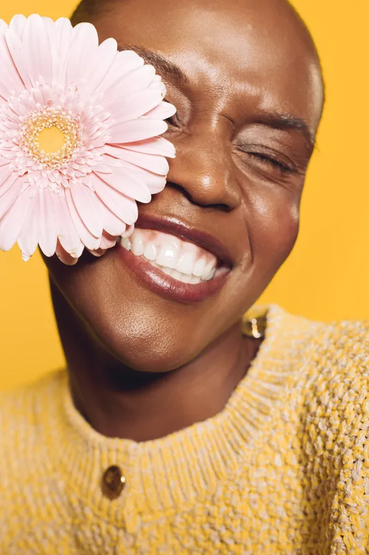 Happy smiling black woman with dark skin holds pink flower near face, eyes closed in joyful expression. Yellow background and sweater create warm, vibrant mood of natural beauty and happiness.