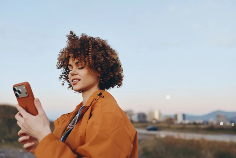 Woman lifestyle portrait, curly hair, orange jacket outdoors, holding smartphone and smiling while looking at screen, casual day scene, distant city skyline and warm soft sky light