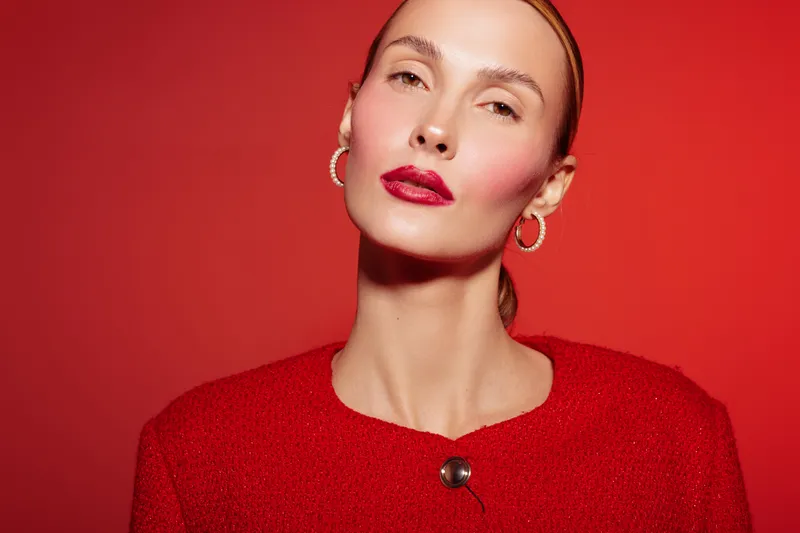 Elegant woman with red lipstick and blush poses against red background wearing red sweater and hoop earrings. Concept of fashion, beauty, makeup, and confident female portrait.