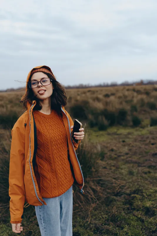Woman outdoors lifestyle walking in a grassy field wearing sweater, jacket, and hat, holding smartphone, wearing glasses, warm casual autumn mood, natural light, countryside background