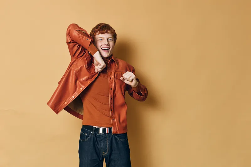 Young man with red hair dancing happily wearing orange shirt and jacket with dark jeans standing against yellow background smiling with energetic movement and casual style.