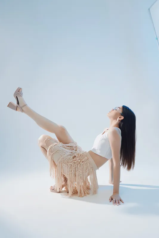 Young woman in a white top and fringed skirt poses with one leg raised in a bright studio. Concept of beauty, fashion, elegance, and modern style with soft natural lighting.