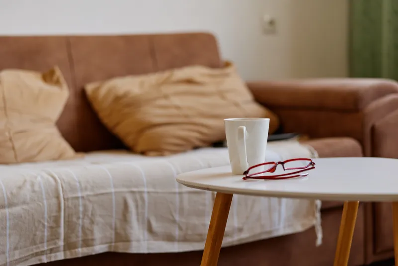 Cozy living room scene shows a white mug on a roundtable beside a sofa, with soft beige cushions and calm natural lighting conveying a relaxed home mood