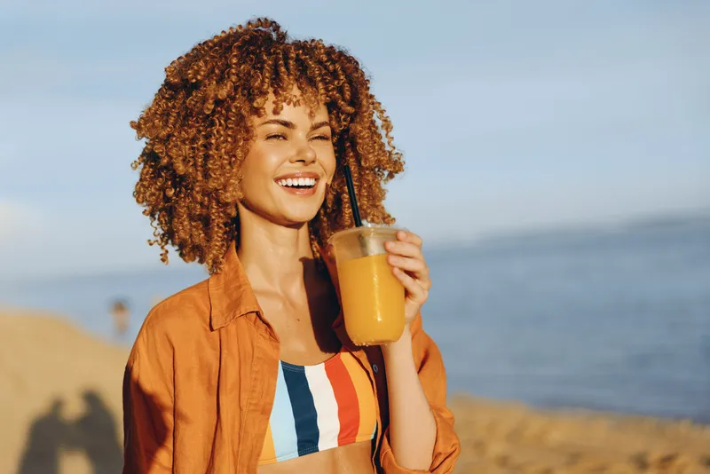 Woman with curly hair smiling, drinking orange juice, casual shirt, standing on beach in summer, enjoying sunlight and sea breeze, fresh wellness lifestyle concept
