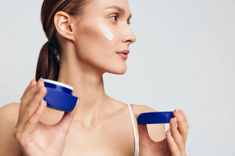 A young woman applies cream from a blue jar on her cheek, standing in a bright studio as she demonstrates a calm skincare moment with a focused, peaceful expression.