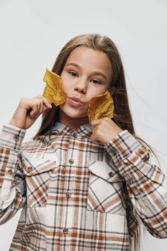 Girl with long hair holding two yellow autumn leaves near face wearing plaid shirt with neutral background. Youth, nature, seasonal and casual clothing concept in studio light.