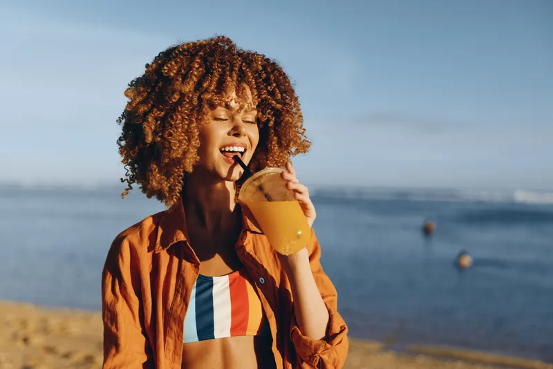 Young woman with curly hair stands on a sunlit beach, sipping juice from a cup, wearing an orange shirt over a striped bikini, radiating joyful summer energy as a light breeze brushes her skin and