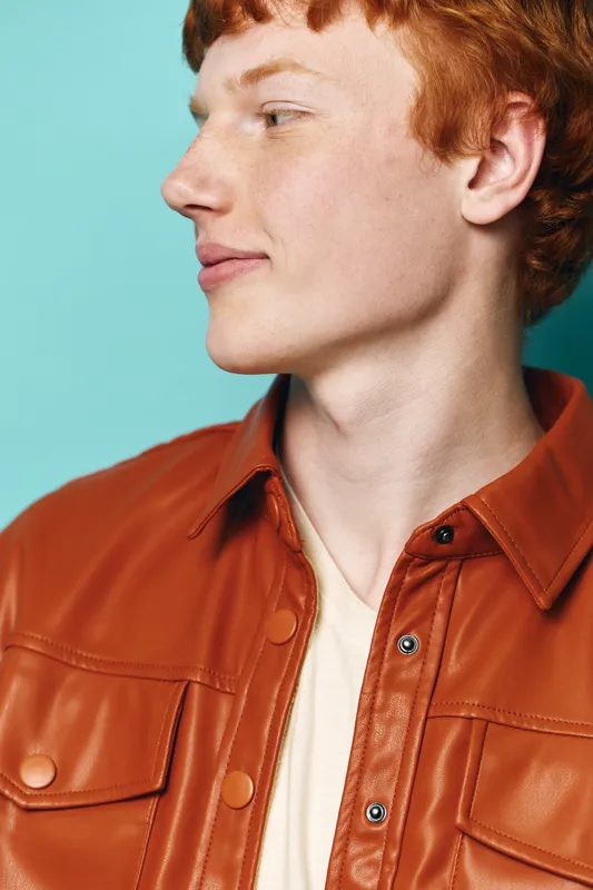 Redhead young man wearing orange leather jacket and white shirt looking sideways with closed lips against blue background in studio portrait with casual style and soft lighting.