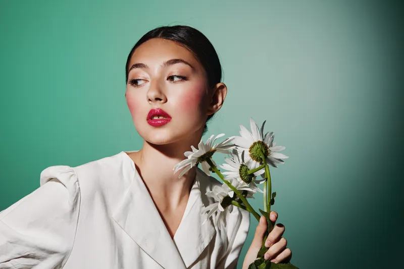 Young woman with glossy red lipstick holding white daisies looks away thoughtfully. Soft light enhances clean fashion styling and beauty creative concept.