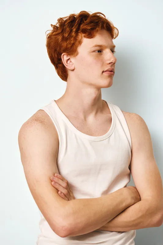 Young man with red hair and light skin wearing white tank top standing with arms crossed looking sideways on plain background. Portrait, casual, confident, and relaxed posture.
