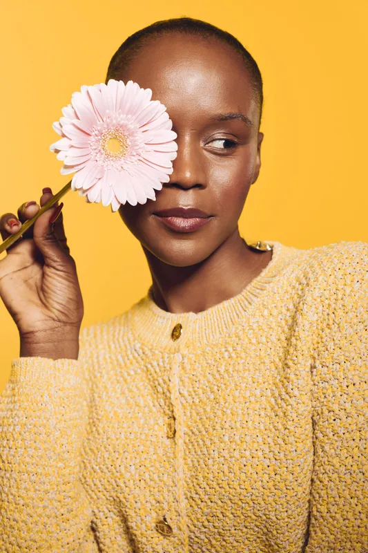 Portrait of a dark skinned African woman holding a pink flower near her eye against a yellow background. She wears a yellow knitted sweater. Concept of beauty, nature, and self-expression.