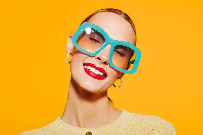 Smiling woman with bright red lipstick wears large turquoise glasses and yellow sweater against orange background. Concept of fashion, beauty, style, and vibrant colors in studio portrait.