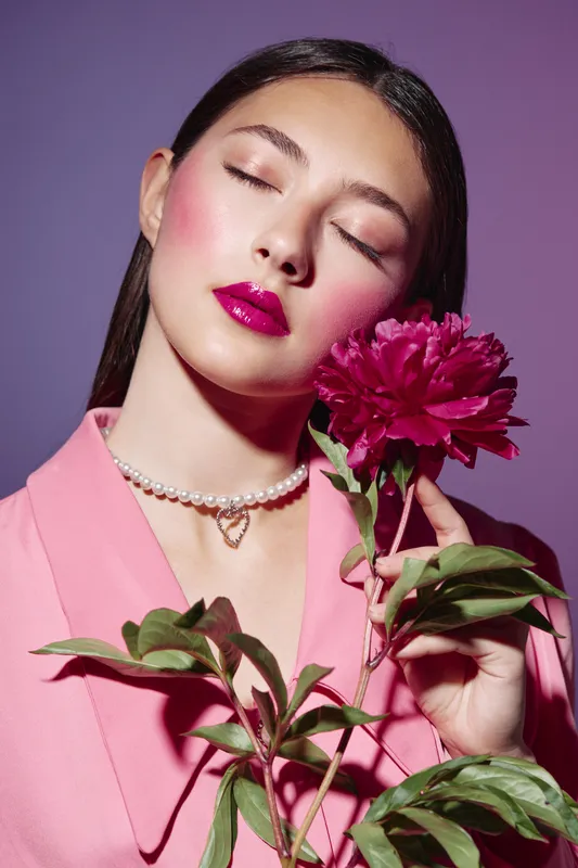 Close-up of a young woman with glossy pink lips wearing a pink blazer and pearl necklace while holding a bright pink flower against a purple backdrop. Beauty creative fashion theme.
