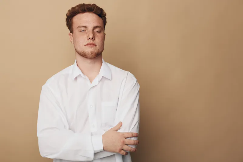 Young man with closed eyes and red hair wearing white shirt stands with crossed arms against beige background. Concept of calm, relaxation, and introspection in casual style.