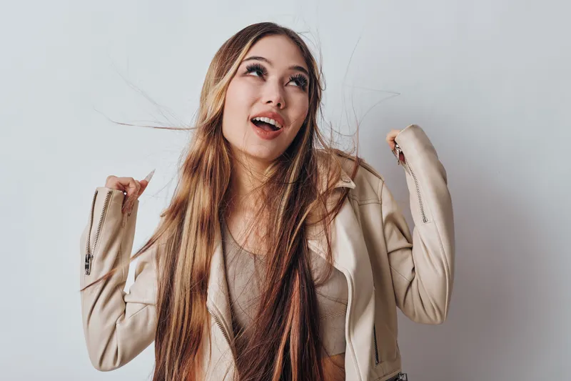Young woman with long hair smiling and raising hands wearing beige jacket and light top posing on white background for lifestyle and happiness concept portrait.