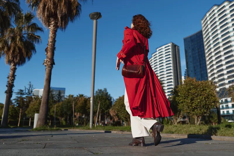 Woman in a red coat walks away along a city park path under blue sky, modern high rises in the background, showcasing confident street fashion and movement