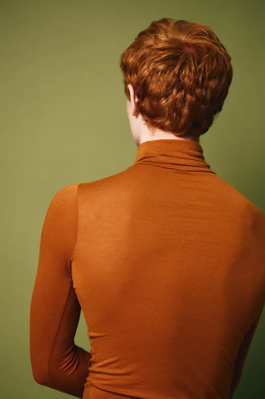 Redhead person standing with back to camera wearing brown turtleneck sweater against green background. Focus on hair and upper body in casual style portrait.