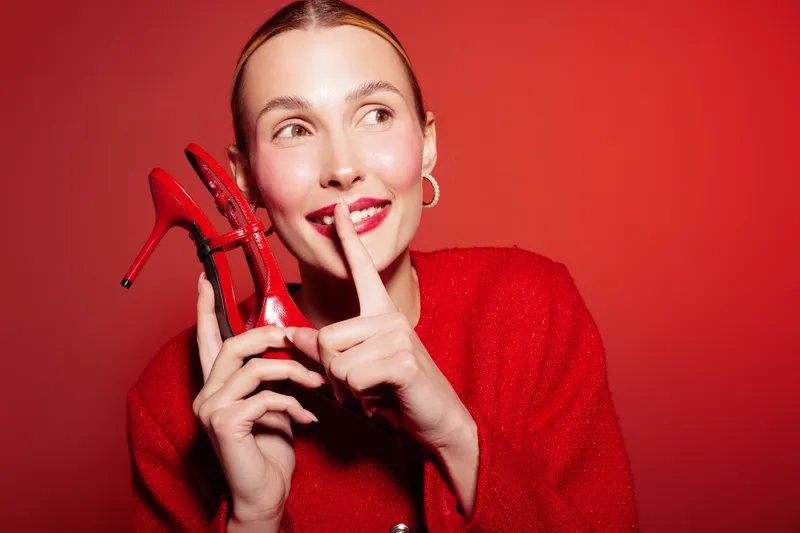 Young woman in red sweater holds red high heel shoes near face, smiling with finger on lips. Concept of fashion, style, beauty, and playful secret on red background.