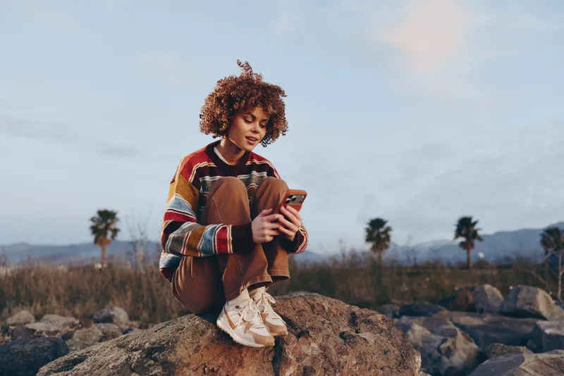 Woman lifestyle outdoors, curly hair, casual style, holding phone on rocky stones, relaxed smile, golden hour sky, distant mountains, natural colors, palm trees