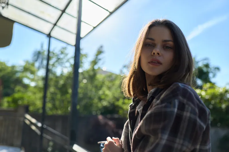 A woman in a plaid shirt stands outdoors in bright daylight, looking over her shoulder with a calm, pensive expression. A lifestyle moment captured in a sunny, urban garden setting, natural