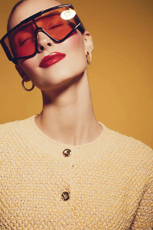 Stylish woman with short hair wears large orange sunglasses and red lipstick. She poses confidently in a textured yellow sweater against a warm yellow background. Fashion concept.