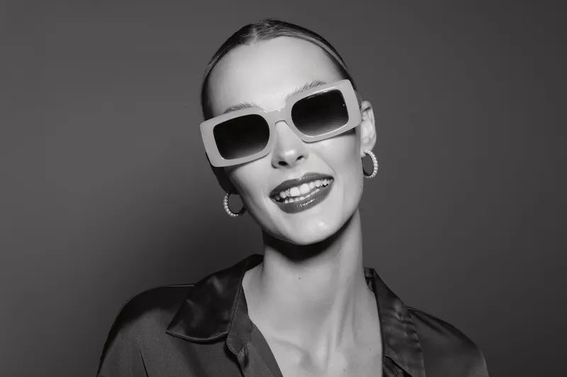 Smiling white woman with pale skin and slicked-back hair wears large sunglasses and hoop earrings in studio. Fashion portrait with confident expression and dark outfit.