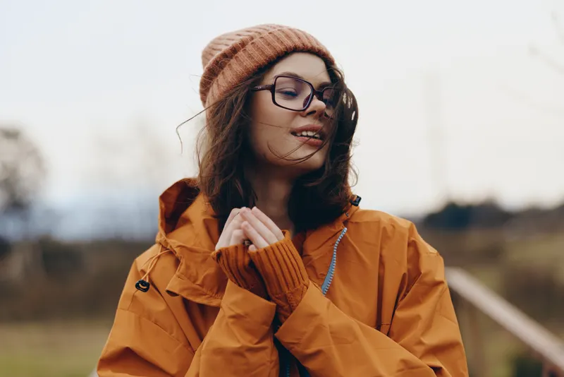 Woman lifestyle outdoors, smiling in an autumn field, wearing an orange coat, knitted beanie, and glasses, soft wind lifting hair, warm natural light, candid portrait, relaxed mood.