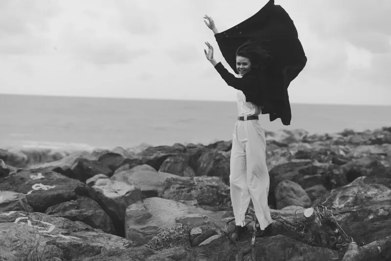 Woman with flowing cape on rocky shore, wind sweeping the sea, capturing motion and freedom in a dramatic monochrome outdoor scene.
