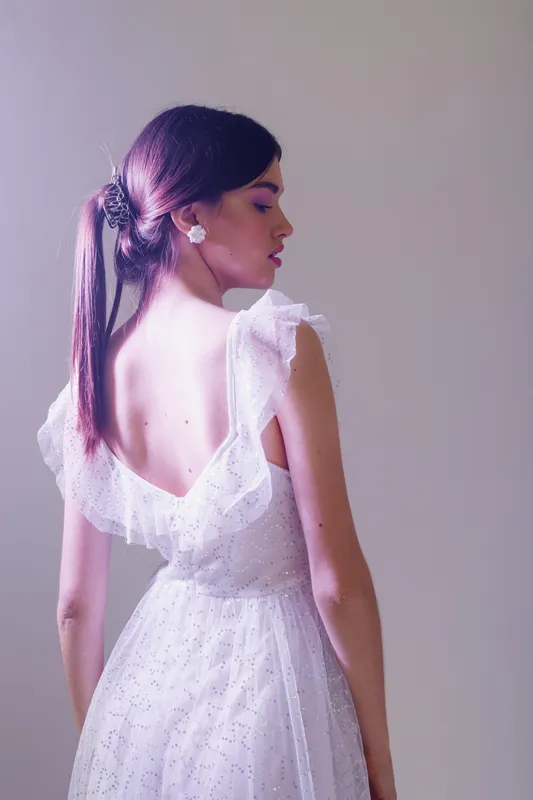 Young woman in a white lace dress with open back poses sideways in soft purple lighting. Concept of beauty, fashion, elegance, and femininity in studio setting.