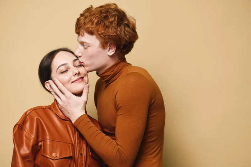 Couple, affection, love and young man kissing woman on cheek with closed eyes. Romantic relationship, emotions and intimacy between people in casual clothing on beige background.