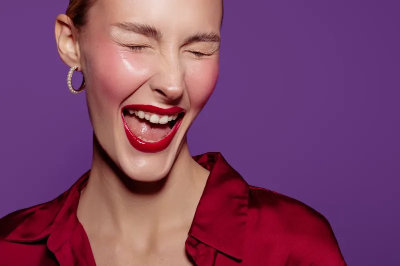 Happy young woman with red lipstick and earrings laughs with eyes closed on purple background. Concept of joy, beauty, fashion, and positive emotions in studio portrait.