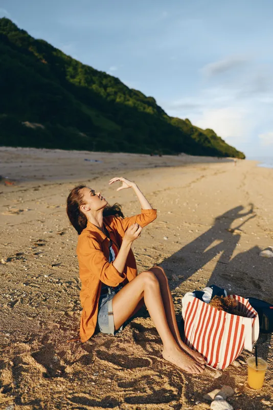 Beach woman sitting on sand in summer vacation, relaxed pose holding phone, golden sunshine and long shadows, striped bag and drink on the beach, travel lifestyle concept, outdoors