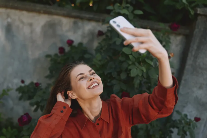 Woman lifestyle selfie in garden, smiling while holding phone to capture sunny moment, casual red shirt, greenery and flowers in frame, relaxed joyful mood, candid portrait